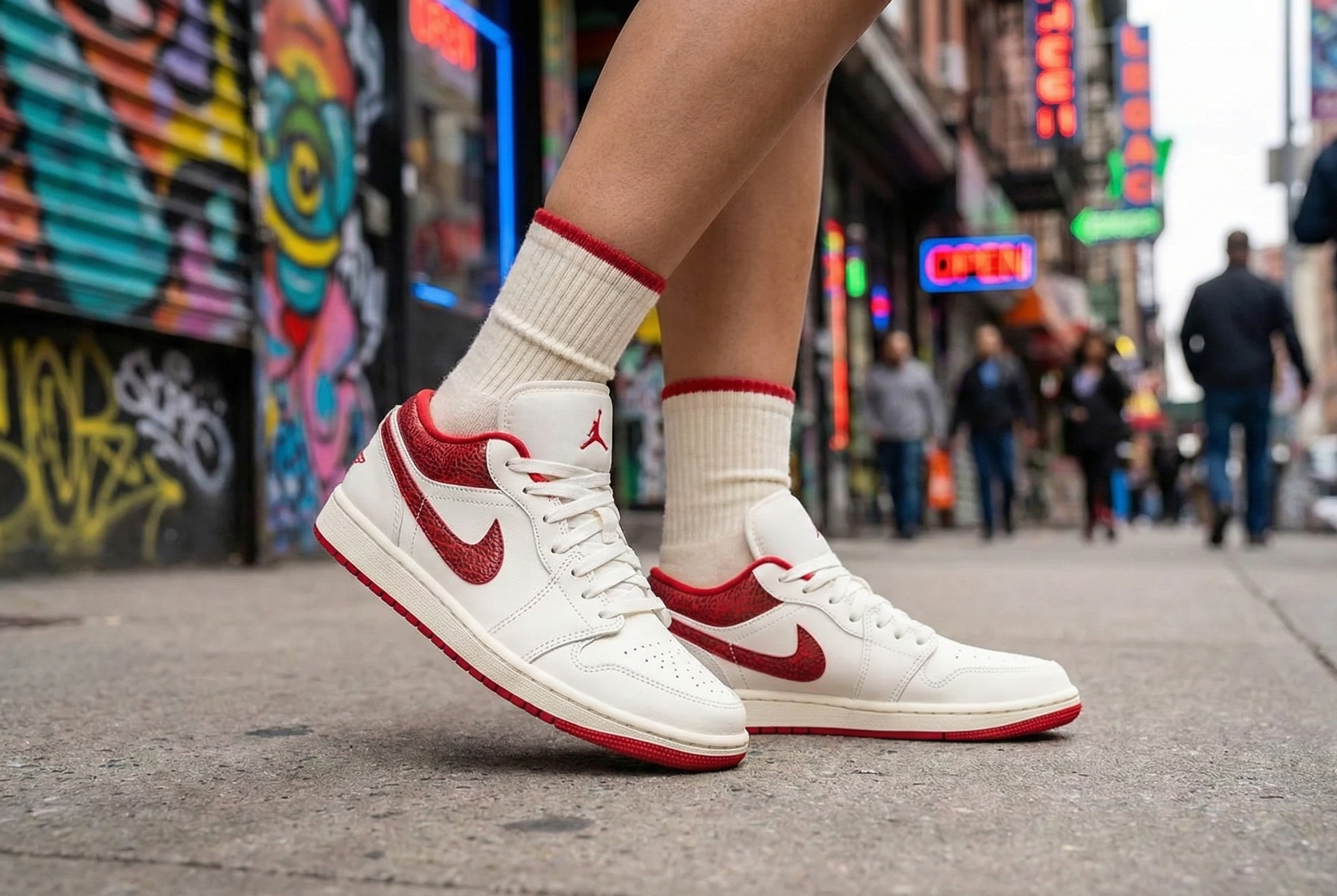 Air Jordan 1 Low SE sneakers with red swooshes worn on a city street with colorful graffiti and neon signs.