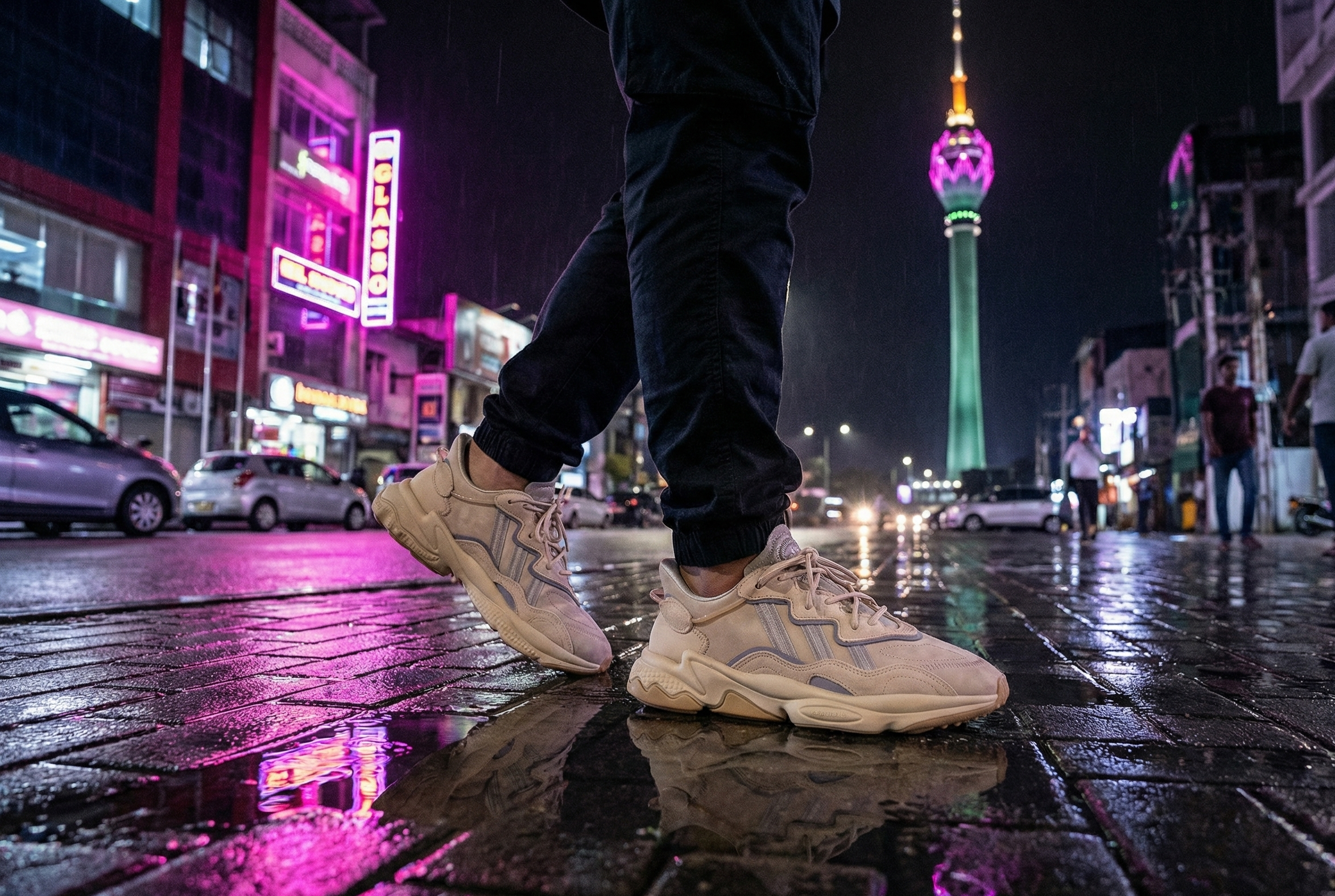 Person wearing beige OZWEEGO Shoes on a wet city street at night with illuminated buildings in Colombo.