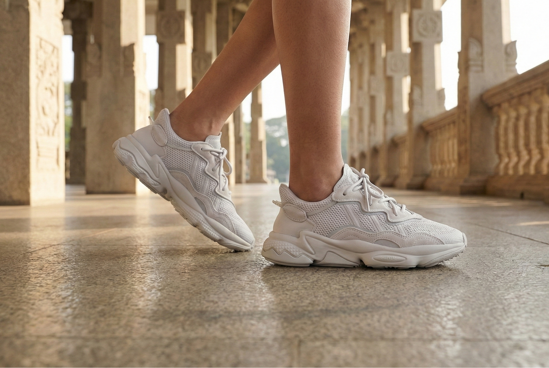 White Adidas OZWEEGO Shoes sneakers worn on a stone floor with an architectural background