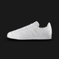 adidas Gazelle shoes in Cloud White and Gold Metallic (Design Code: BB5498). Featuring a premium full-grain leather upper in a tonal Triple White colorway with matching white three-stripes and tongue. Finished with a durable rubber cupsole and iconic Gold Metallic "GAZELLE" branding on the quarter panel. Luxuntique offers 100% authentic adidas leather heritage sneakers at the lowest price in Sri Lanka.