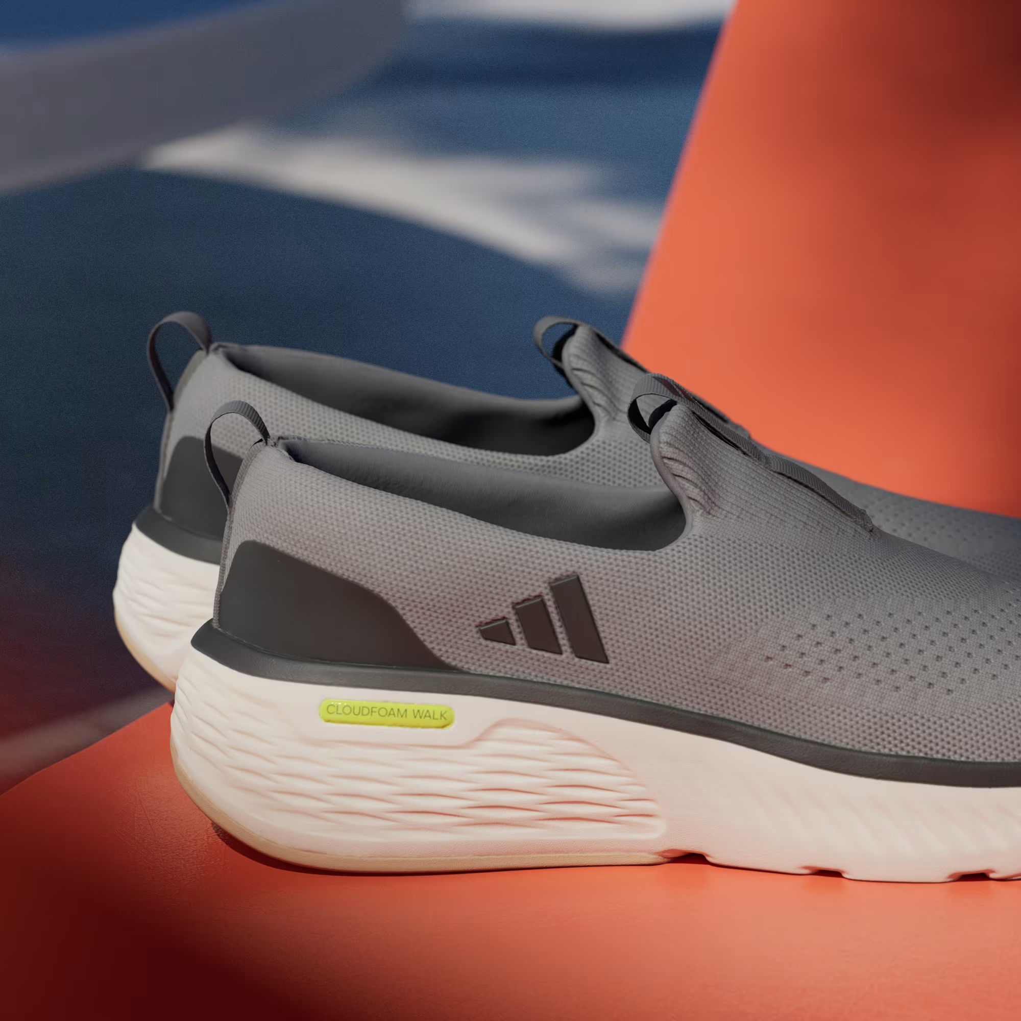 Adidas Cloudfoam Go Lounger ID4021 - Heel Close-up on Orange Surface showing Grey Three Knit Mesh Texture, Dark Grey Three Stripes, Dual Pull Tabs, Neon Yellow Cloudfoam Walk Midsole Branding & Off White Chunky Midsole