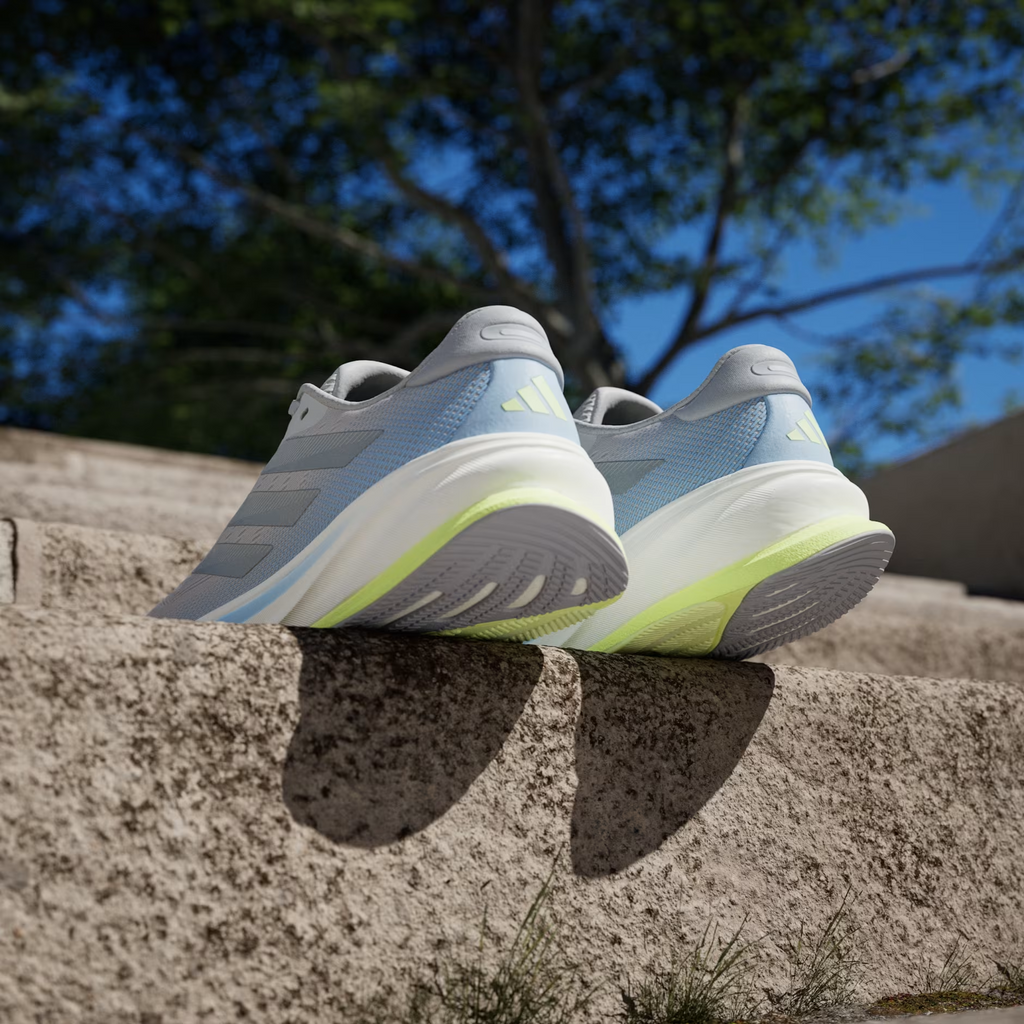 Adidas Supernova Rise 2 IH8708 - Front 3/4 Pair Shot on Stone Ledge with Urban Background showing Grey Two Mesh Upper, Tonal Matte Silver Three Stripes, Hi-Res Yellow Tongue Badge, Light Blue Midsole Accent & White Dreamstrike+ Midsole