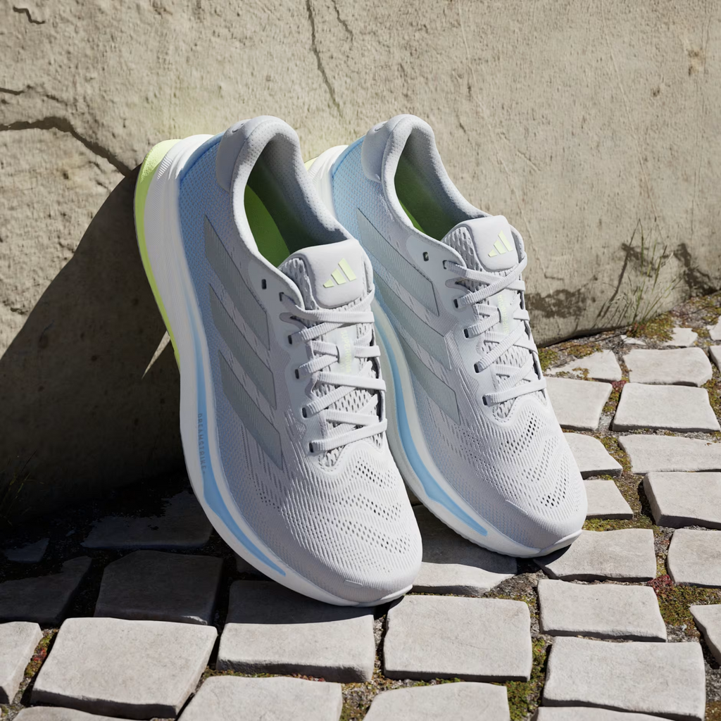 Adidas Supernova Rise 2 IH8708 - Clean Lateral Profile on Cobblestone Ground with Bokeh Background showing Full Grey Two Silhouette, Tonal Matte Silver Three Stripes, Light Blue Midsole Stripe, White Dreamstrike+ Midsole & Hi-Res Yellow Outsole