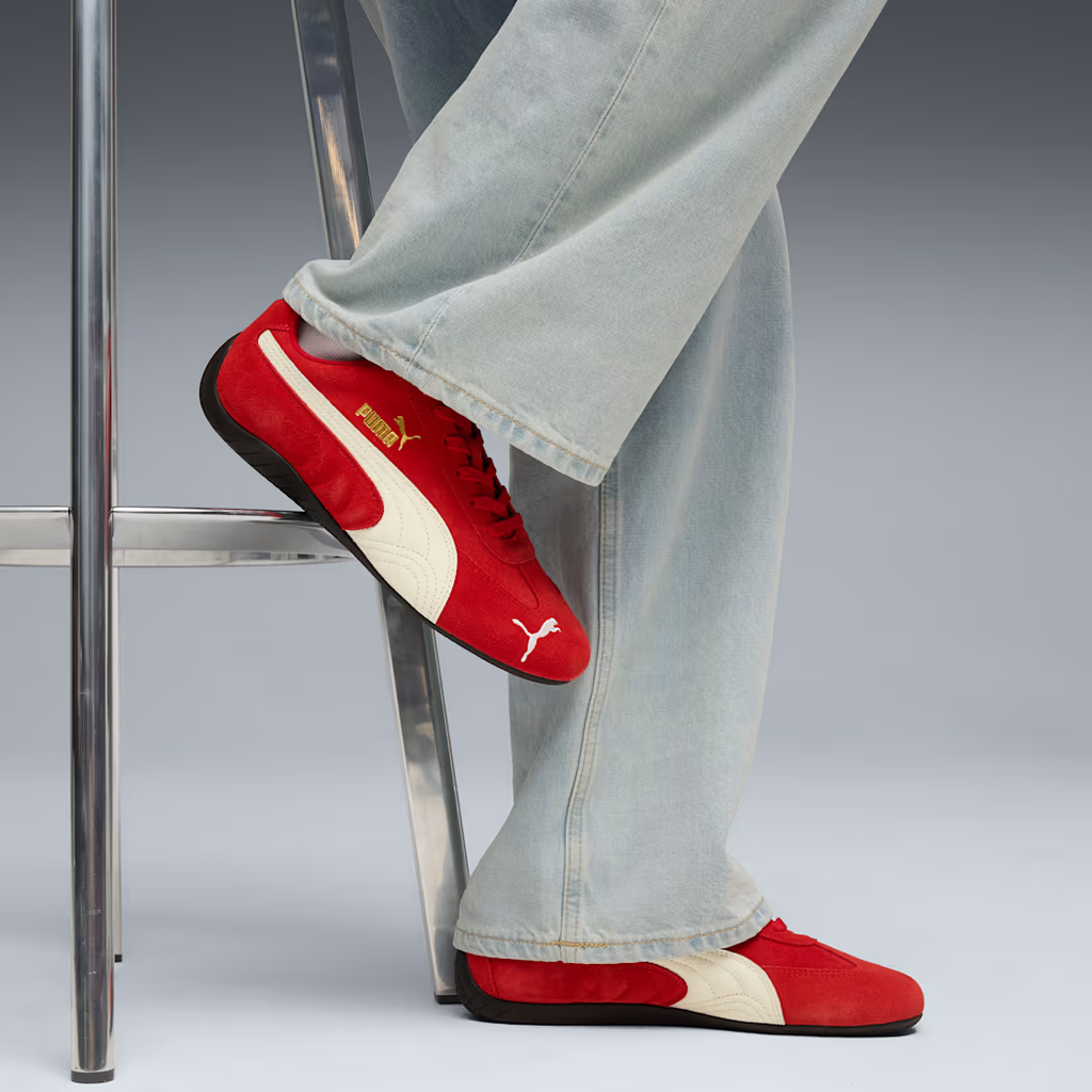 PUMA Speedcat 406329_02 - On-Foot Lifestyle Shot on Chrome Chair with Grey Studio Background showing For All Time Red Suede Upper, PUMA White Formstripe, Gold PUMA Wordmark, Red Flat Laces & Black Rubber Outsole worn with Light Wash Wide-Leg Jeans