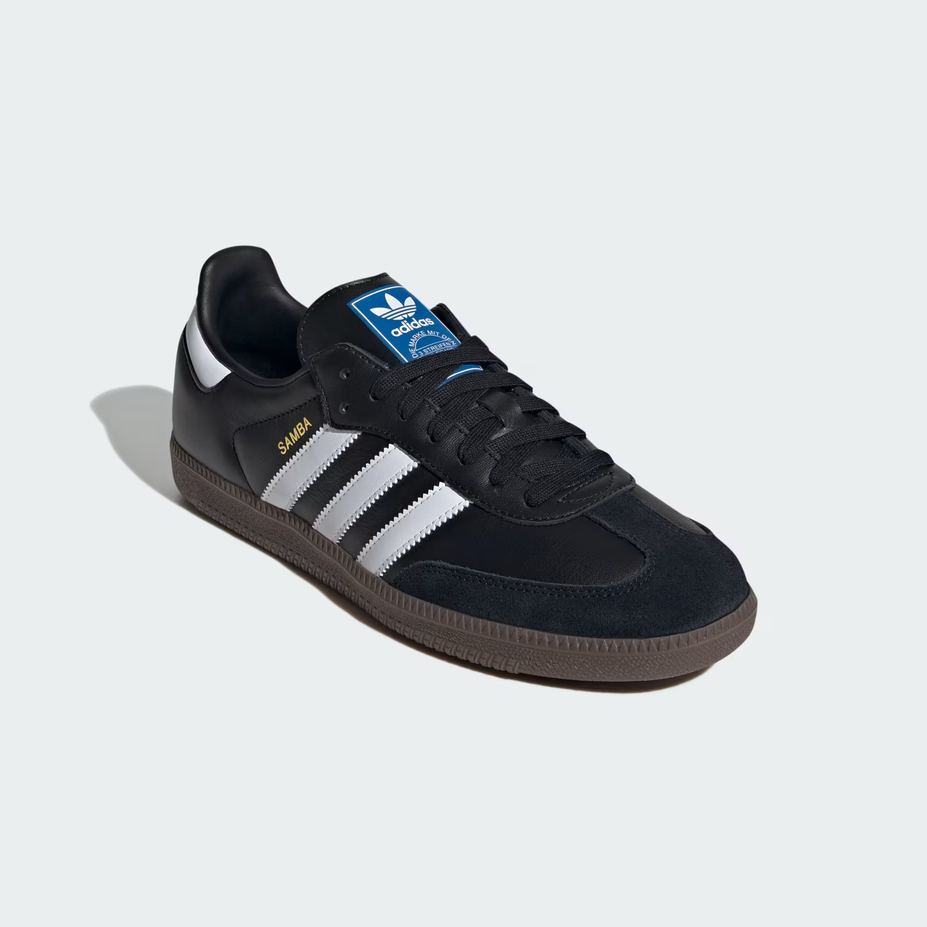 Adidas Samba OG B75807 - Lateral Profile Lifestyle Shot on Light Grey Background with Loose Black Laces showing Core Black Leather Upper, Gold Samba Wordmark, Cloud White Three Stripes, Black Suede Toe Box, Blue Adidas Trefoil Tongue Badge & Gum Rubber Outsole